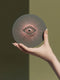 Round ceramic coaster featuring a sage green background with a detailed rose gold Evil Eye symbol in the center, surrounded by radiating metallic rays in a sunburst pattern.